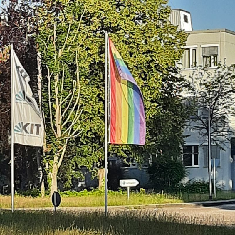 Zwei Fahnen im Freien: Regenbogenfahne und KIT-Fahne vor einem Gebäude.